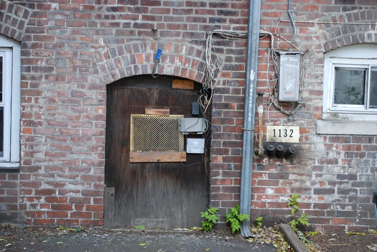 A worn wooden plank door under a brick arch, with a vent grille fitted into it and utility boxes on the wall beside it