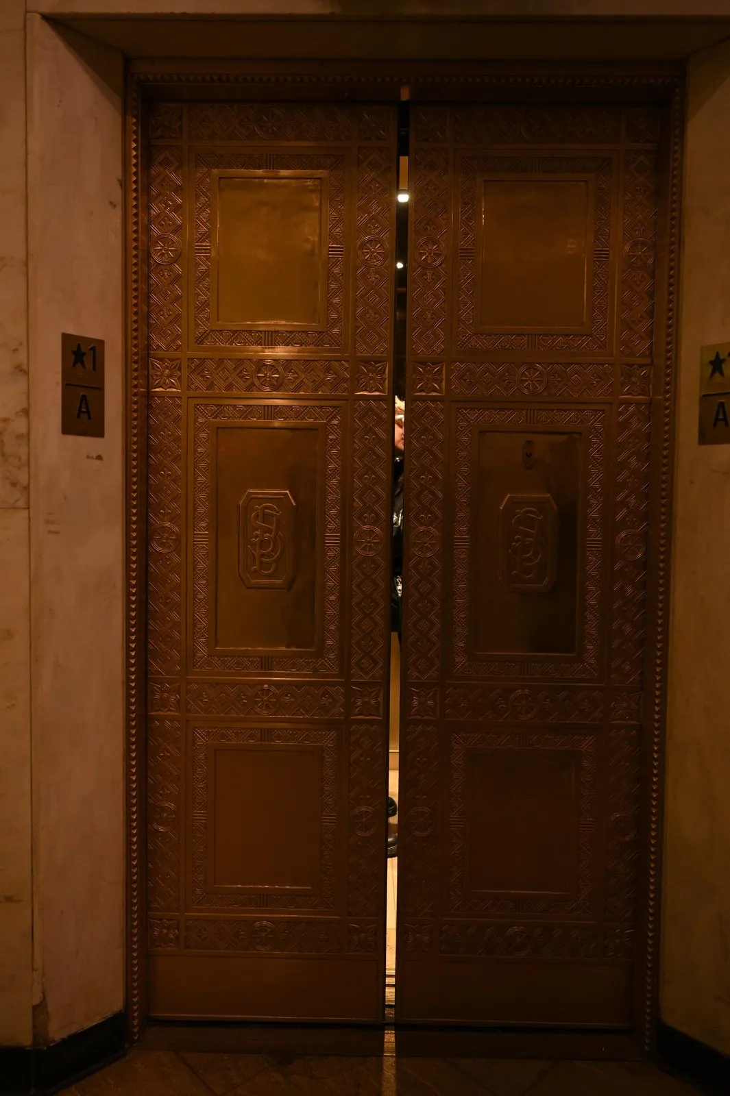 Paired embossed metal elevator doors, slightly parted, glowing gold in a dim hallway