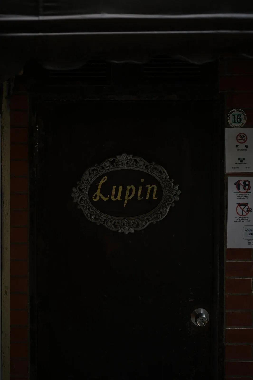 A dark door with an oval scrollwork nameplate reading Lupin
